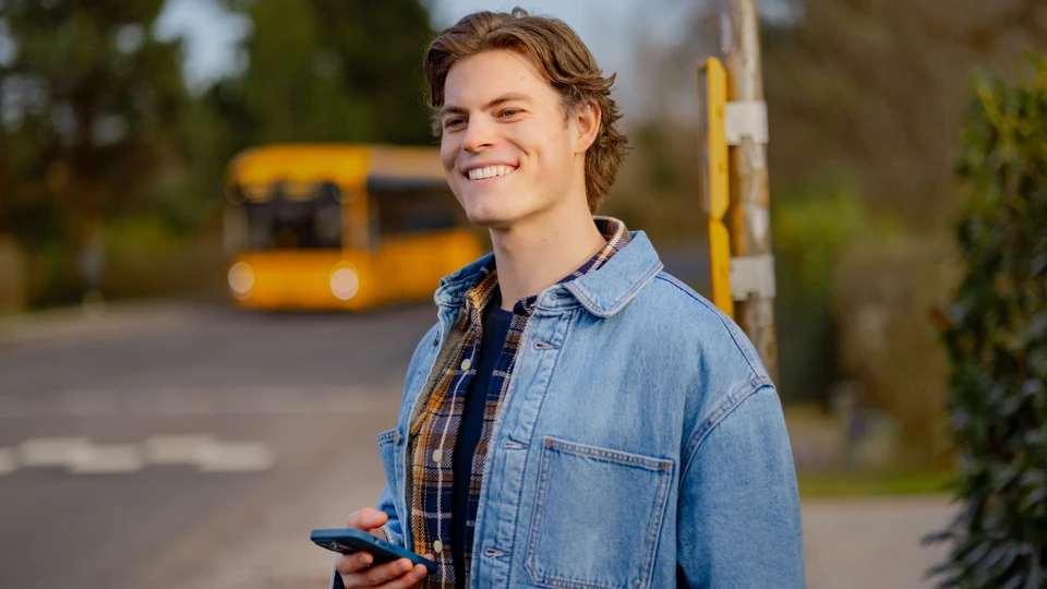 En smilende ung mand med sin telefon, der venter på bussen